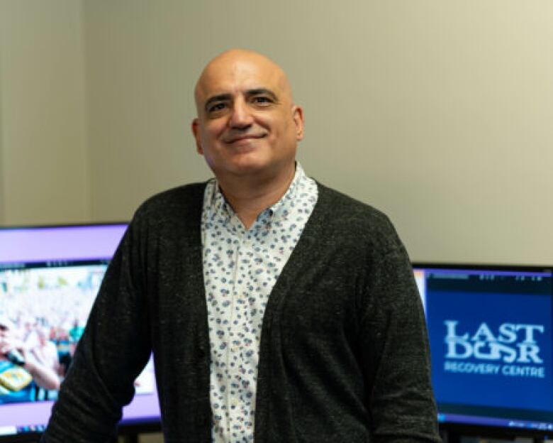 A bald man in a white shirt and dark cardigan smiles at the camera. He is standing in an office.