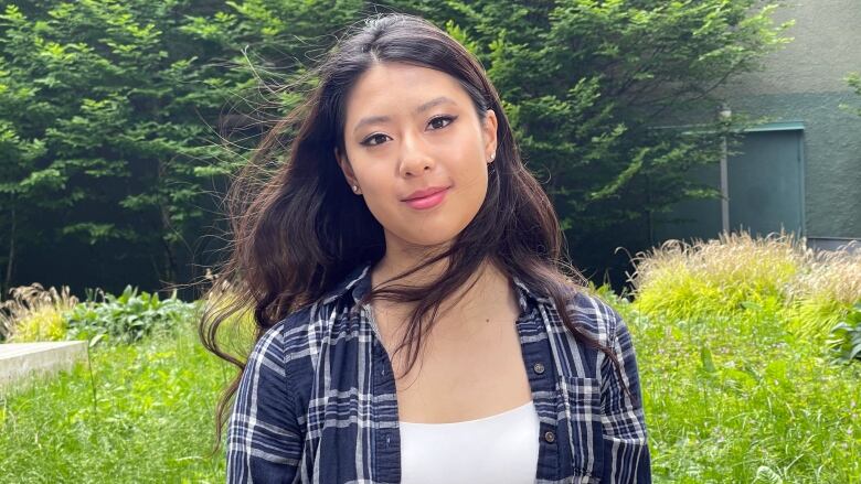 A young East Asian woman with long black hair smiles in the sunshine in front of a grassy field. She is wearing a blue plaid shirt over a white tank top and her hair is blowing in the wind.