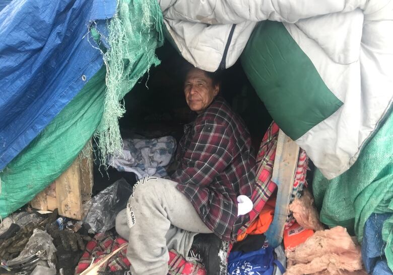 A man squats at the entrance of a tent wearing a black and red plaid shirt and grey sweatpants 