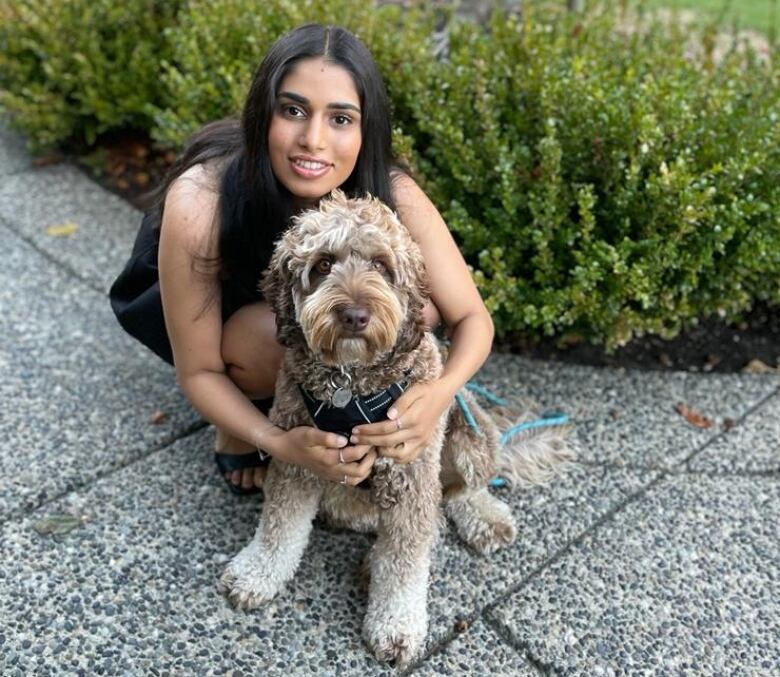 A young South Asian woman with long, dark hair crouches down on a sidewalk to hug a shaggy dog.