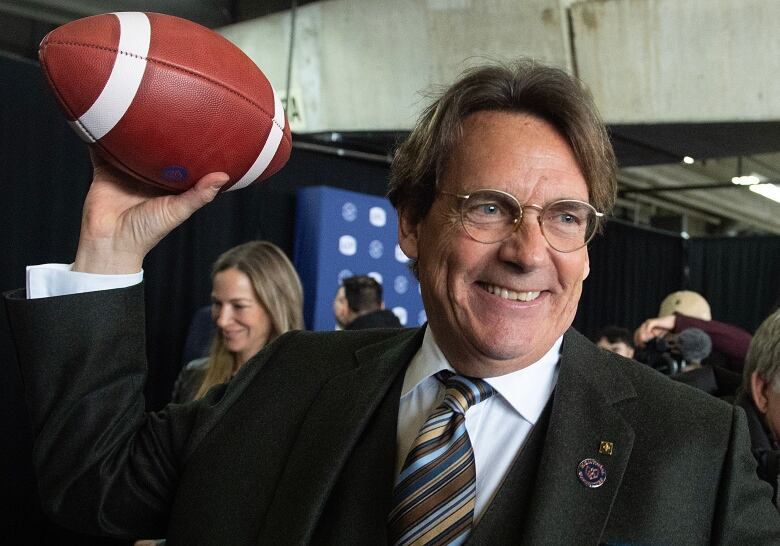 Smiling man in a suit hoists a football in his right hand.