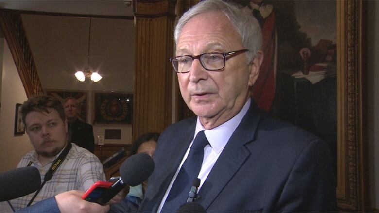 A man with grey hair and glasses, wearing a dark suit, white shirt and tie, speaks to reporters.