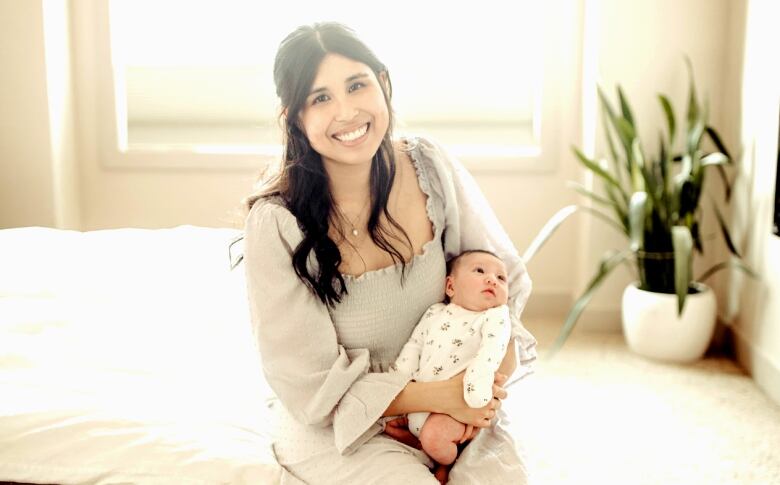 A woman wearing a white dress sits on a bed holding a baby.