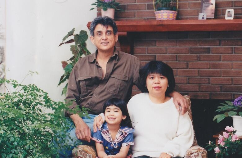 A young girl and a woman sit in an arm chair while a man sits on the arm of the chair and all smile for a posed family photograph. 