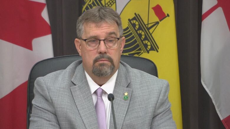 A man with glasses wearing a grey suit sits behind a desk with flags behind him.