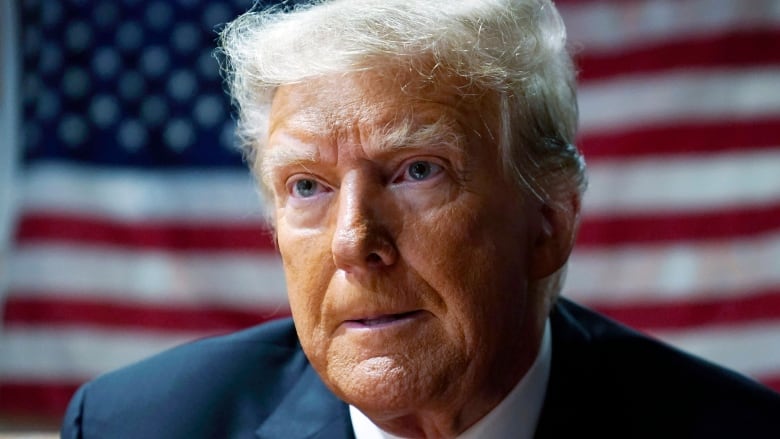 A close-up image shows Donald Trump, wearing a dark suit and a red tie, looking past the camera while sitting in front of an American flag. 