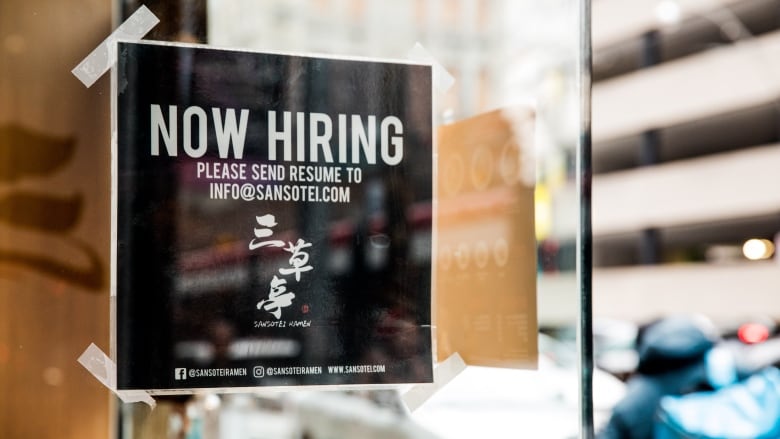 A 'now hiring' sign is taped to the glass door of a restaurant.