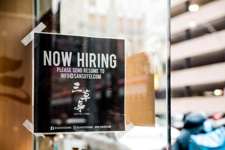 A 'now hiring' sign is taped to the glass door of a restaurant.