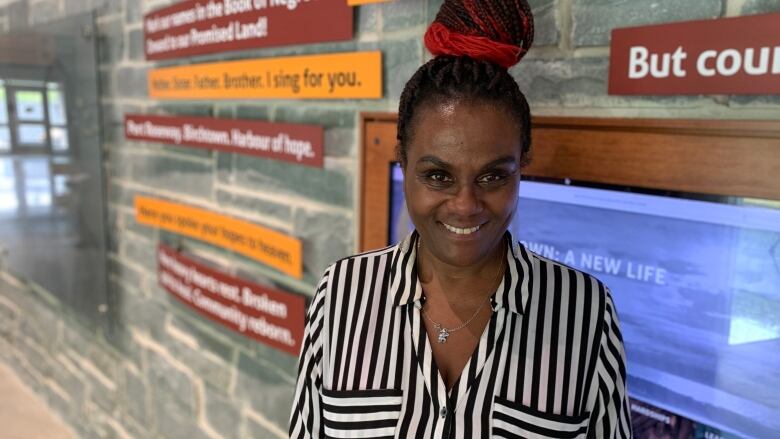 A woman wearing a black-and-white striped shirt smiles as she stands in front of a stone wall adorned with signs and a television.