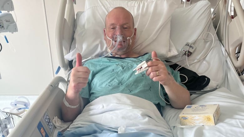 A man wears an oxygen mask and a blue hospital gown. He is laying in a hospital bed and giving two thumbs up. 