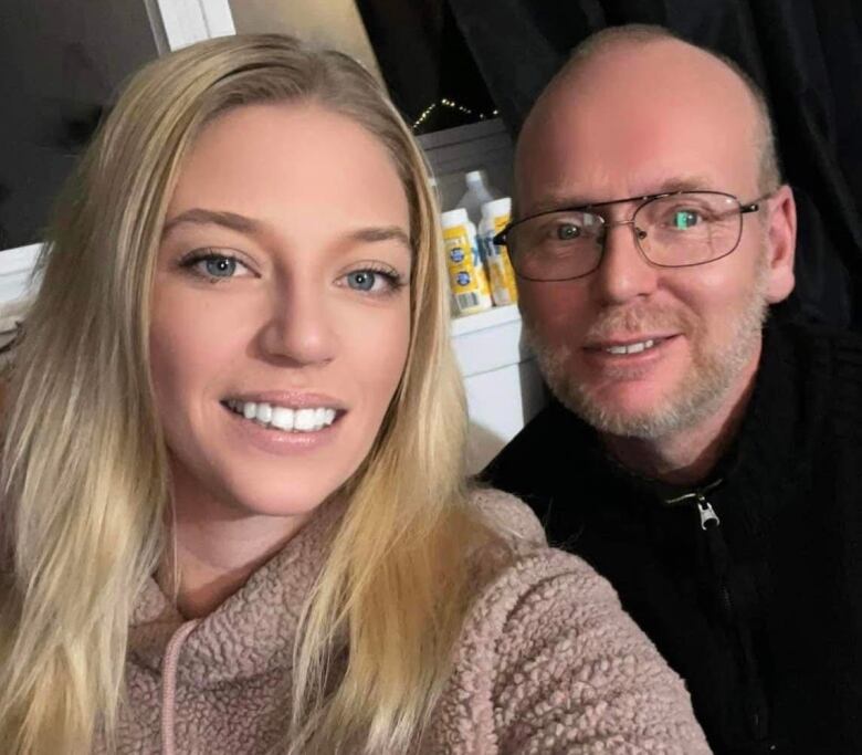 A woman with blonde hair smiles next to an older man who is also smiling and wearing glasses. 