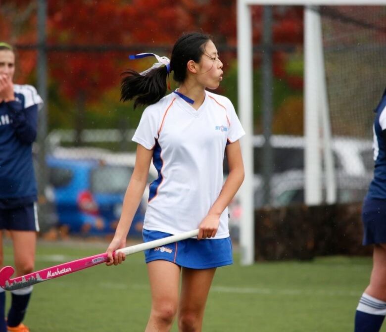 An East Asian teenage girl wearing a blue and white sports uniform holds a pink field hockey stick during a game. Two other players can be seen in the background.