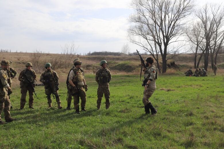 A person in a military uniform holding a weapon talks to seven other people wearing military uniforms as they stand in a field.