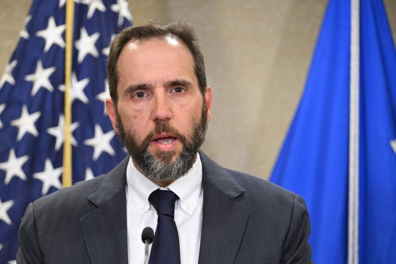 A man with a beard, wearing a dark grey suit and black tie, is shown mid-speech. Behind him is the red, white and blue American flag.