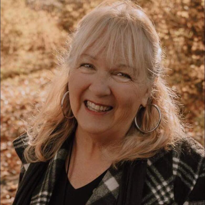 A blond woman wearing silver hoop earrings and a black and white plaid jacket smiles.  