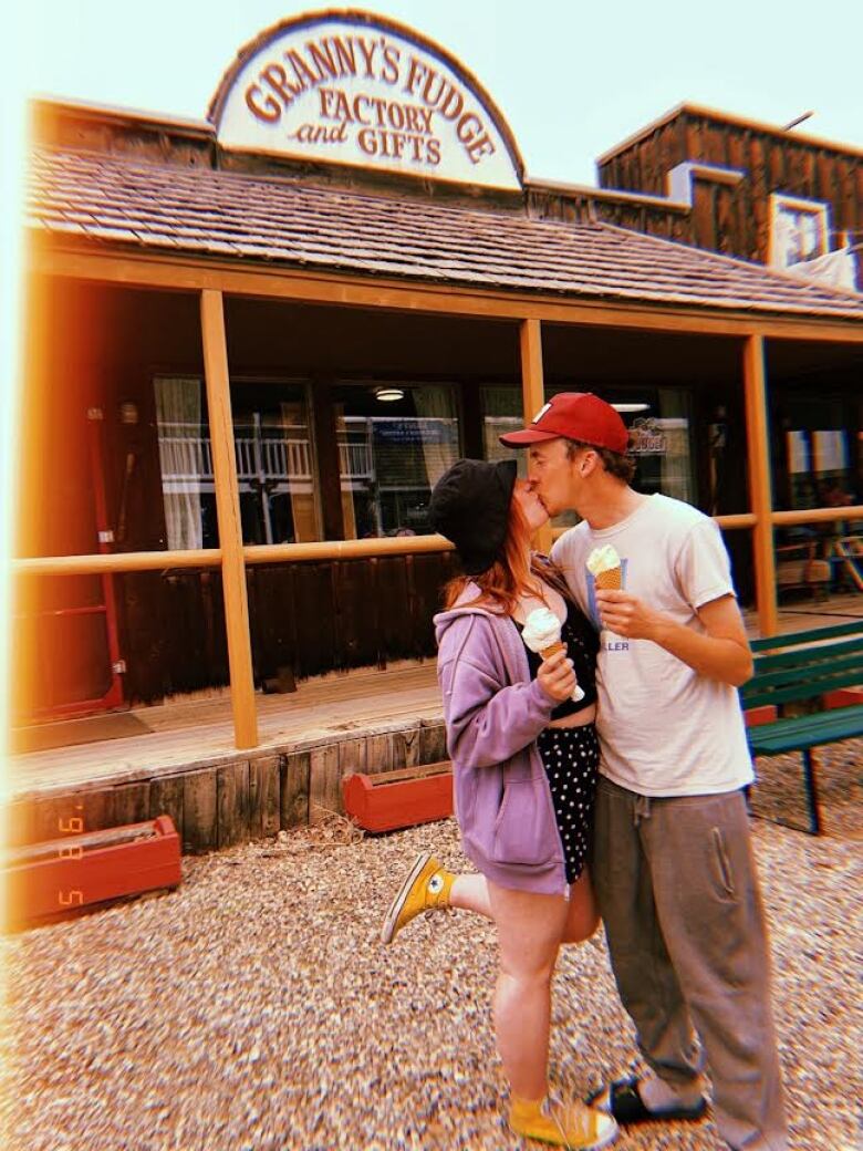 A couple holding ice cream cones kisses in front of a building. 