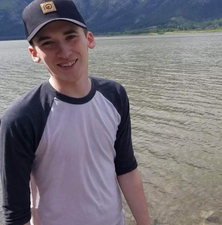 A young man wearing a baseball shirt smiles in front of a lake. 