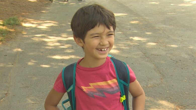 Young boy with red shirt and a blue backpack smiles with a small gap in his teeth. 