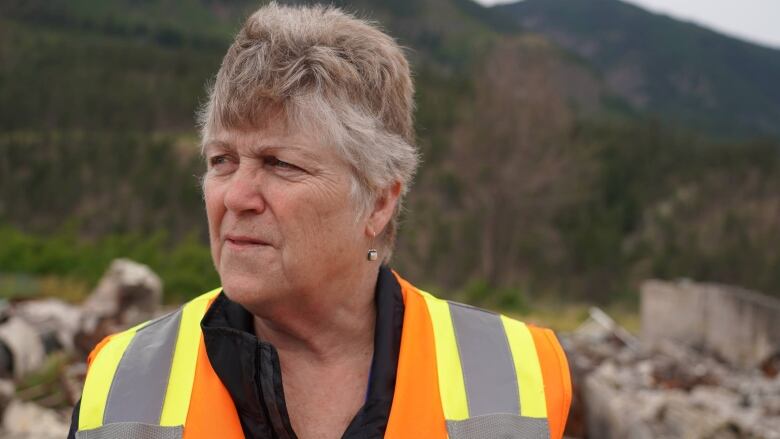 A woman with short brown and white hair in a high-vis vest stands among rubble.