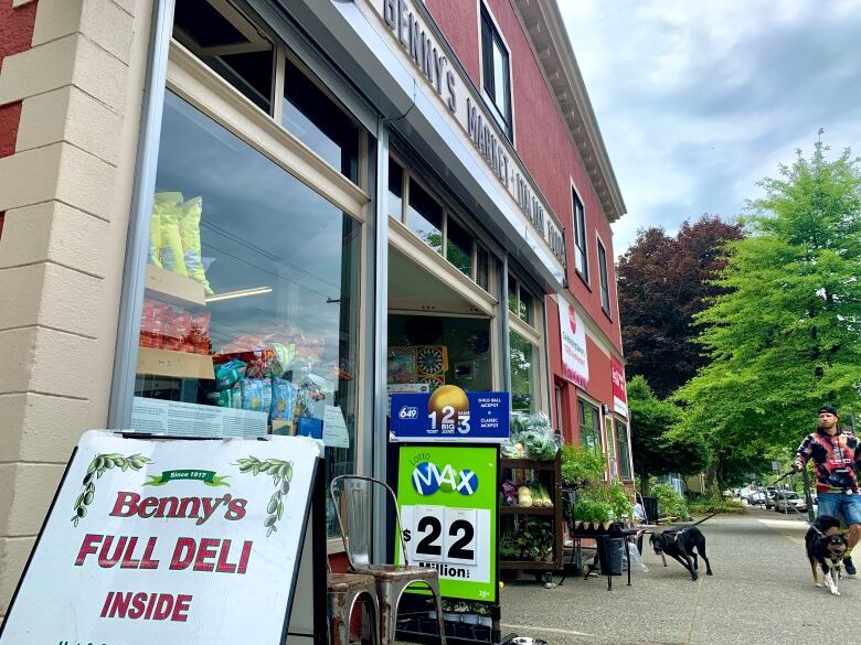 A sign says Benny's Full Deli Inside outside a shop selling plants, produce, Italian food and lottery tickets, as a man walks his two dogs past.