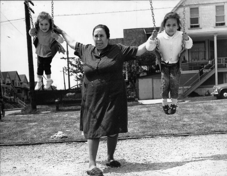 A woman pushes two children on play ground swing set with a house behind them.