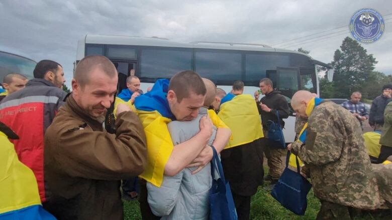 A man with a blue and yellow Ukrainian flag draped over his shoulder hugs another person with other people crowded in the background in front of a bus. 