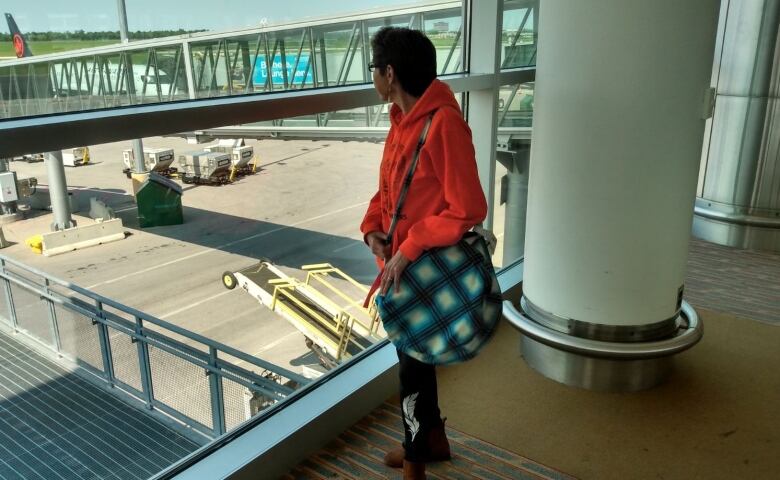 A woman in an orange hoodie looks out a window at an airport.