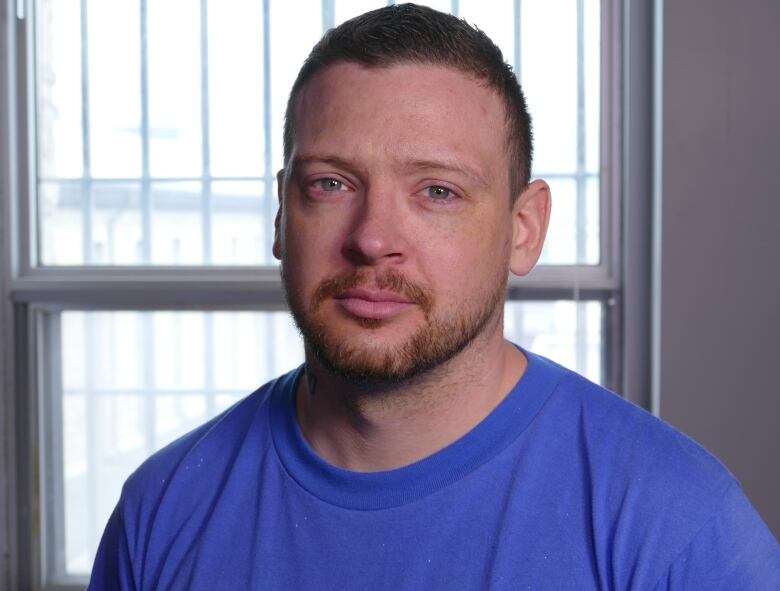 A man with short light brown hair and wearing a blue T-shirt stares into the camera, in front of a window with metal bars on it.