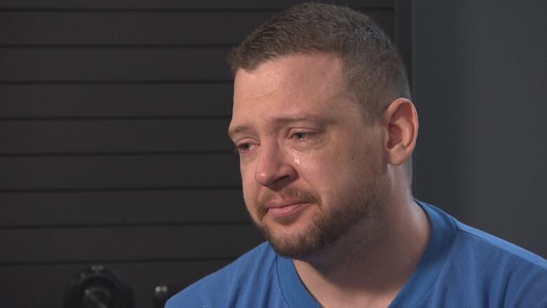 A man with light brown hair and wearing a blue t-shirt stares off to the side, as a tear rolls down his face.