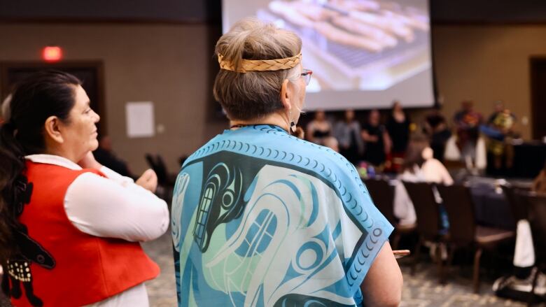 Two women are seen from the back standing in a meeting room with people visible and a projector screen visible in the background.