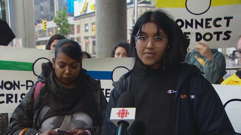 A woman speaks into a microphone while protestors hold signs behind her.