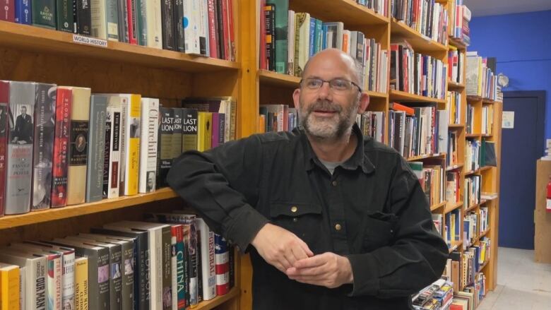 A man with glasses stands, smiling, leaning on a bookshelf in a bookstore. 