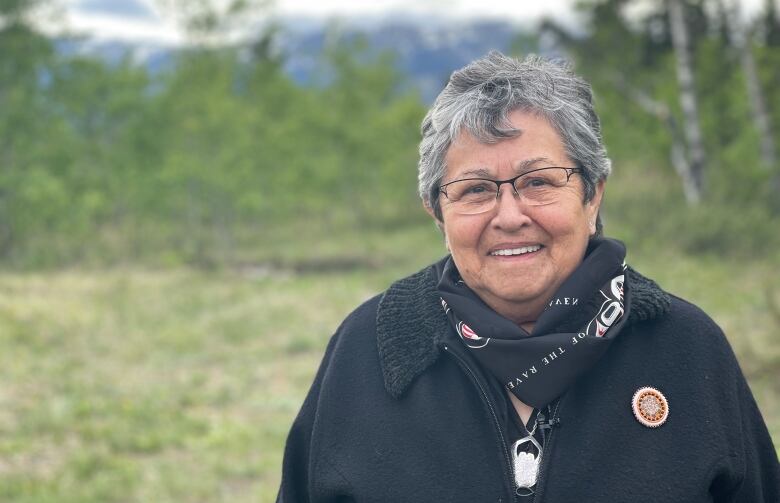 A woman with short grey-black hair, wearing a black jacket and glasses, smiles as she poses for a portrait outside.