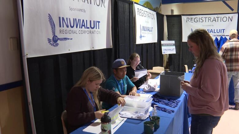 A woman stands before a table where three people sit in front of a sign for the Inuvialuit Regional Corporation, at a trade show.