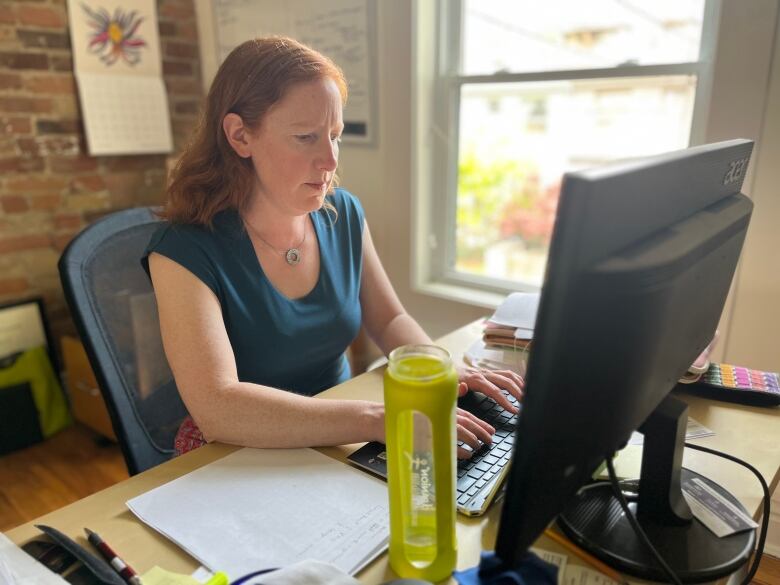 A woman with long red hair sits at a desk facing a computer monitor with her hands on top of the keyboard. Behind her is a brick wall and a window.