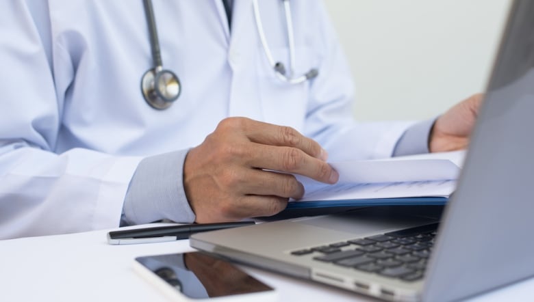 Doctor reading patient health record chart and working on laptop computer on white desk in hospital with copy space, electronic health records system EHRs, teleconference or telemedicine concept.