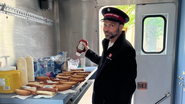 A man in a salvation army uniform prepares hot dogs inside a food cart.