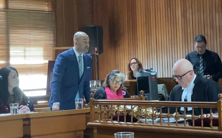 A man in a suit stands speaking in a room of people sitting at tables and desks.