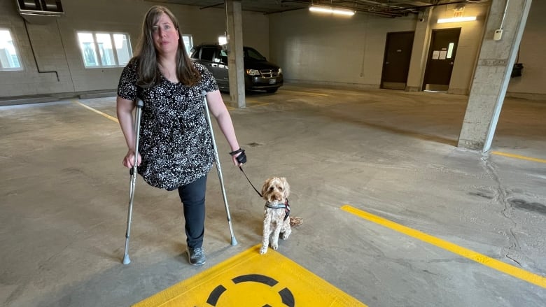 A woman with one leg amputated uses crutches and stands in an indoor parking garage holding a dog on a leash. 