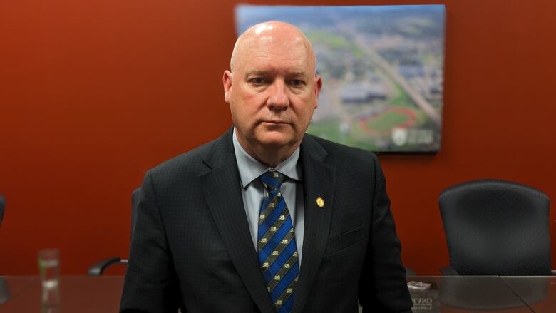 Man with serious expression stands in an office, wearing a business suit.