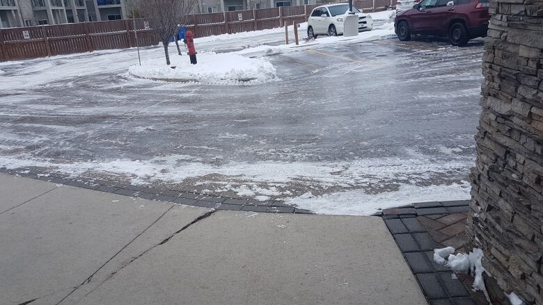 A parking lot outside an apartment building is covered with ice, with a fire hydrant and a white car in the background. 