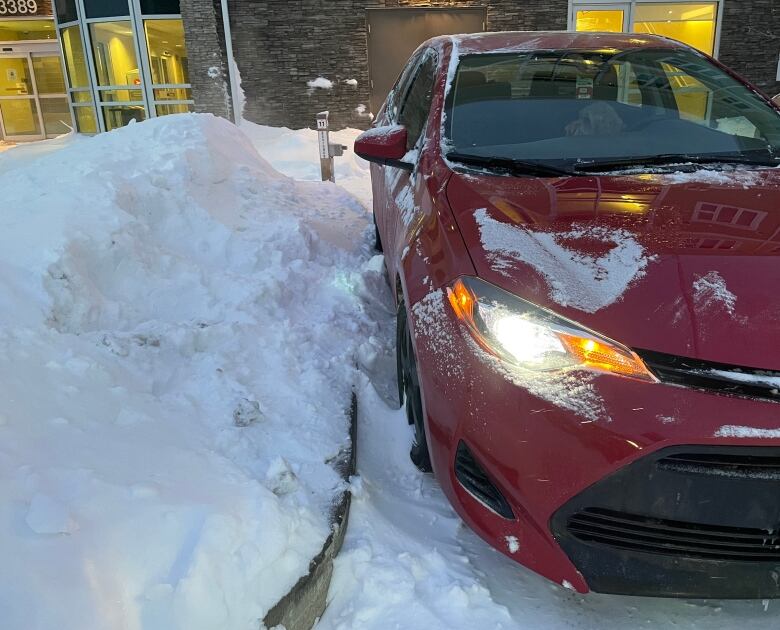 A snowbank is in the space beside a red car parked on a lot outside an apartment building.