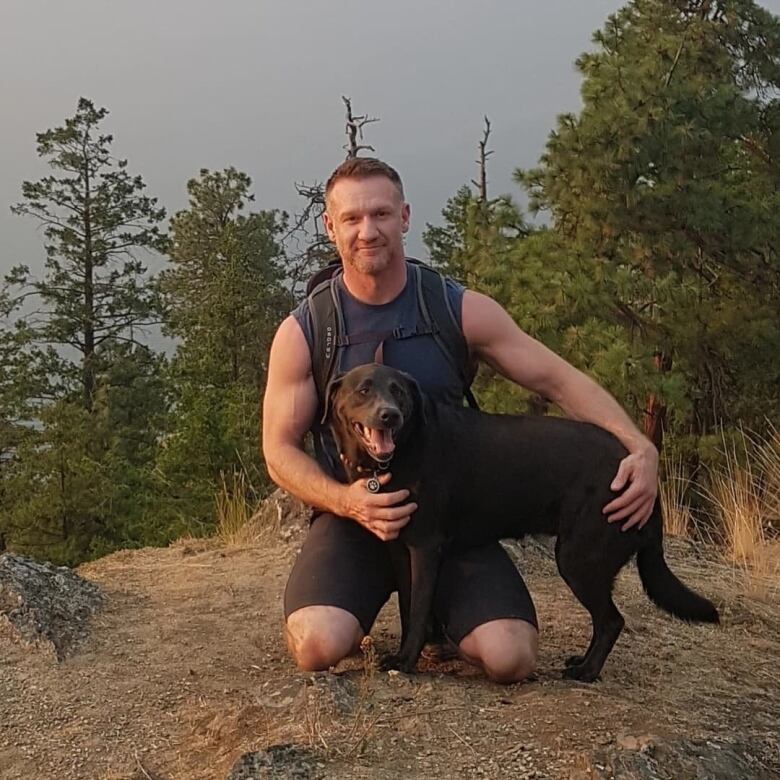 A man kneels down near his dog as they perch at the top of a hill