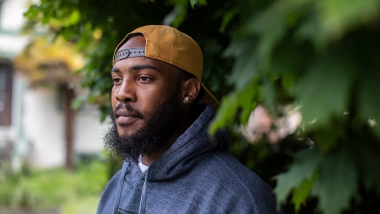 A Black man in a blue hoodie and yellow ball cap is pictured standing next to a tree in a garden.