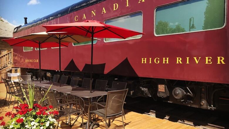 A red train that reads 'High River' in yellow text on the side. Out front is patio furniture and flowers. 