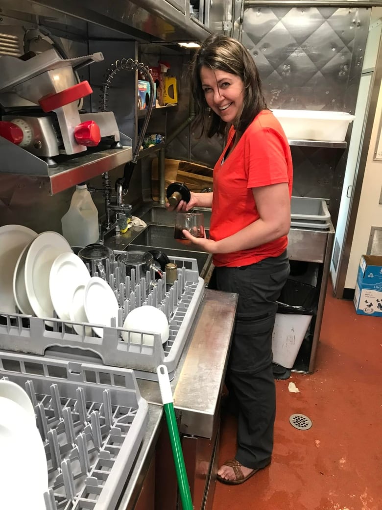 A woman in a red shirt smiles while doing dishes. She is wearing sandals. 