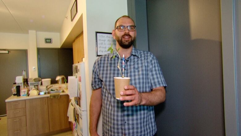 A man with glasses stands in an apartment holding a paper cup with a plant growing in it.