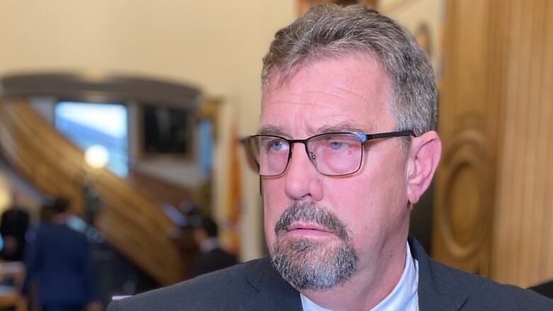 A man wearing glasses and grey suit talks to reporters in a hallway.