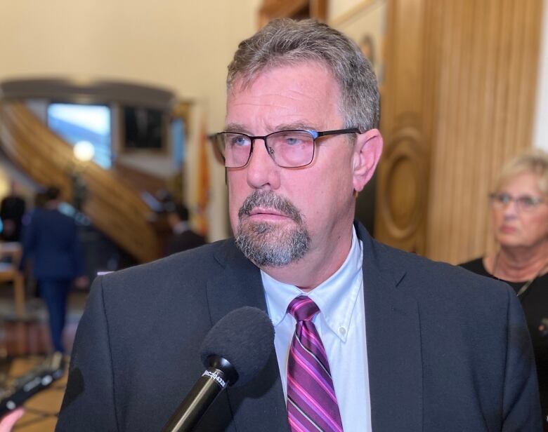 A man wearing glasses and grey suit talks to reporters in a hallway.