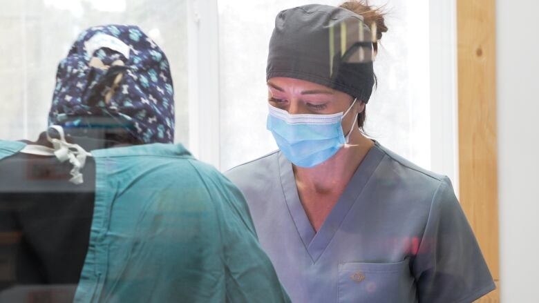 A woman in a smock and cap wearing a mask, assisted by another woman in a green smock, looks down.